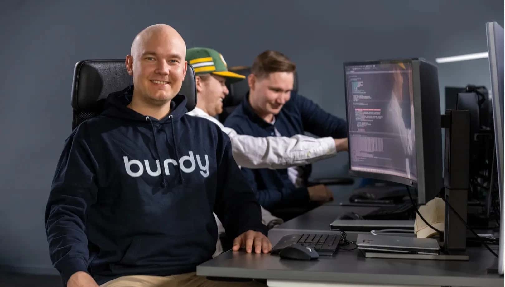 A male employee sits on a computer chair with a hoodie with the text "Burdy" facing the camera. Besides him he has his computer and behind him his two colleagues are point to another computer screen